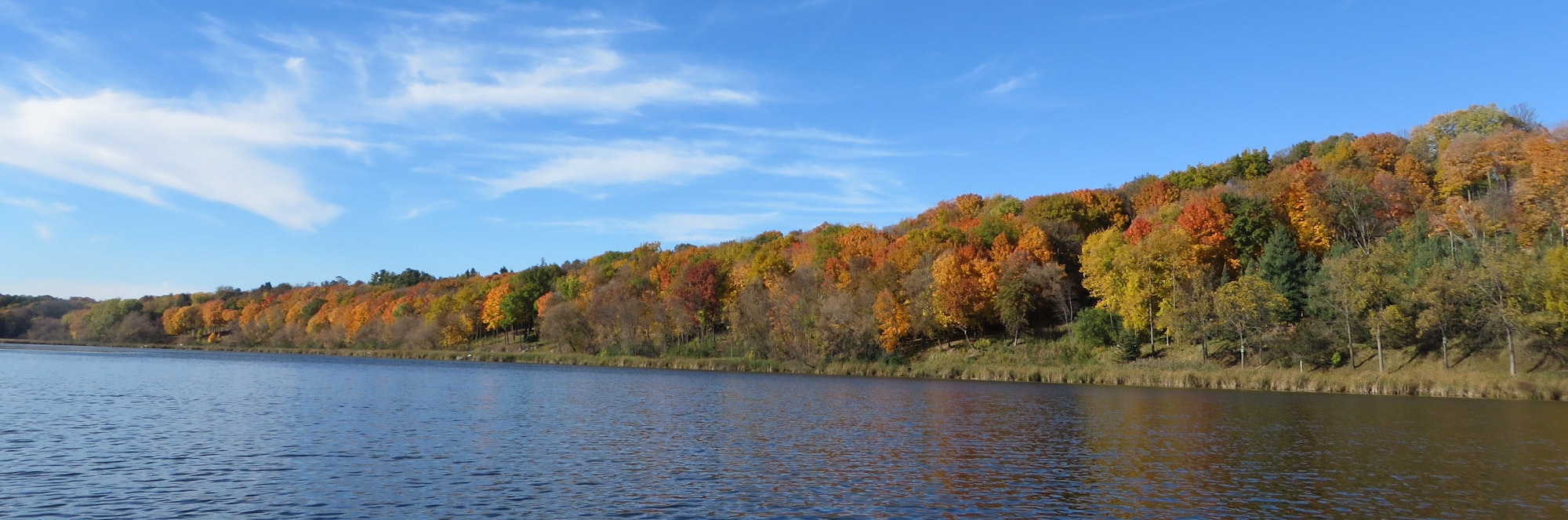Autumn foliage on a hillside by a lake under a blue sky with wispy clouds.
