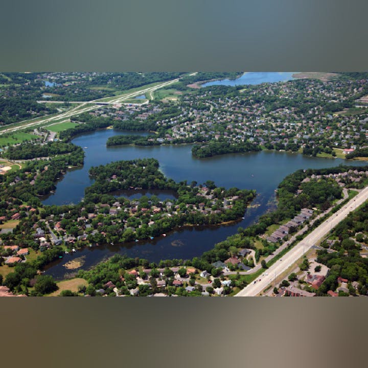 Aerial view of a suburban area with a lake, houses, trees, roads, and some open land.
