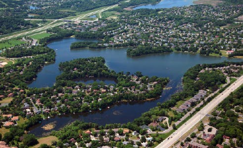 Aerial view of a suburban area with a lake, houses, trees, roads, and some open land.