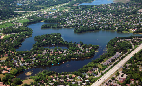 Aerial view of a suburban area with a lake, houses, trees, roads, and some open land.