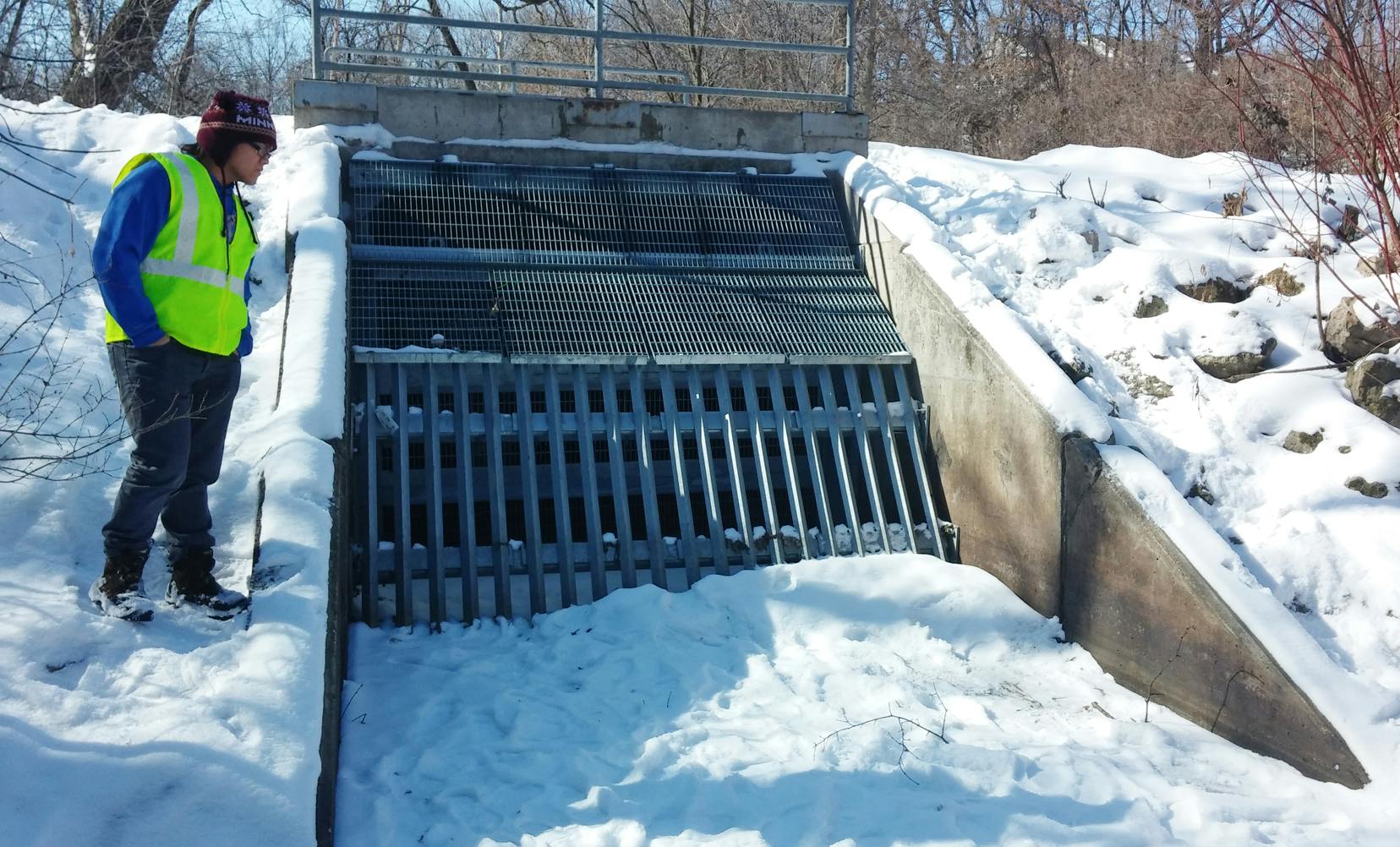A person in a hi-vis vest by a snow-covered metal grate barrier, likely a flood control structure.