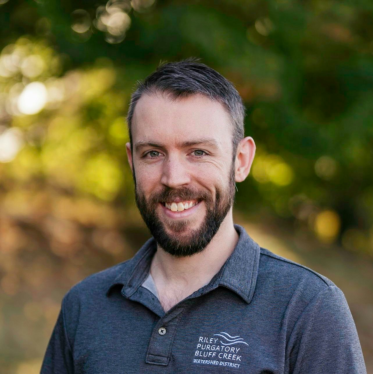A man with a beard smiles outdoors, wearing a "Riley Purgatory Bluff Creek Watershed District" polo.
