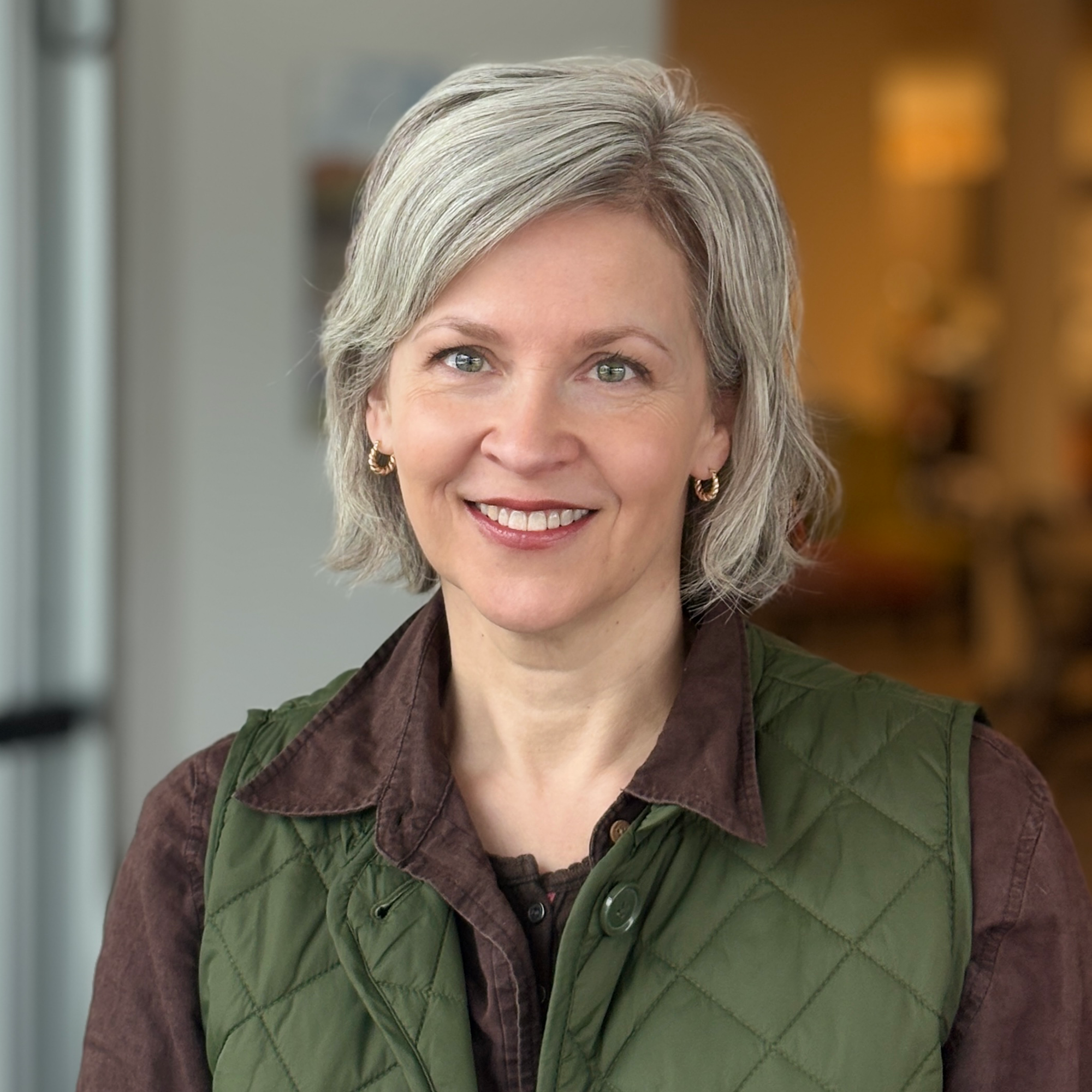 A woman with short, gray hair smiles warmly, wearing a green quilted vest over a brown shirt, in a bright indoor setting.