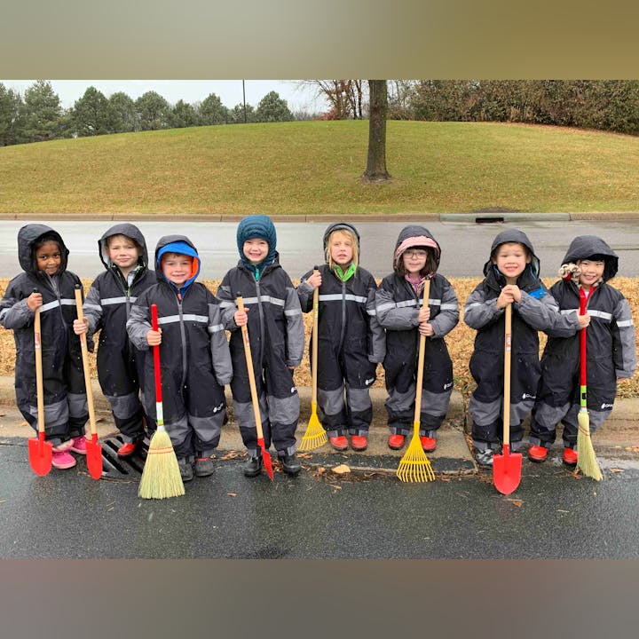Group of children in rain gear holding garden tools, standing on a street on a rainy day.