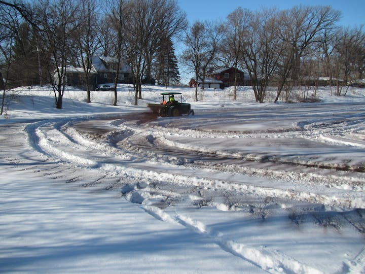 A tractor in a snowy landscape with tire tracks and houses in the background.