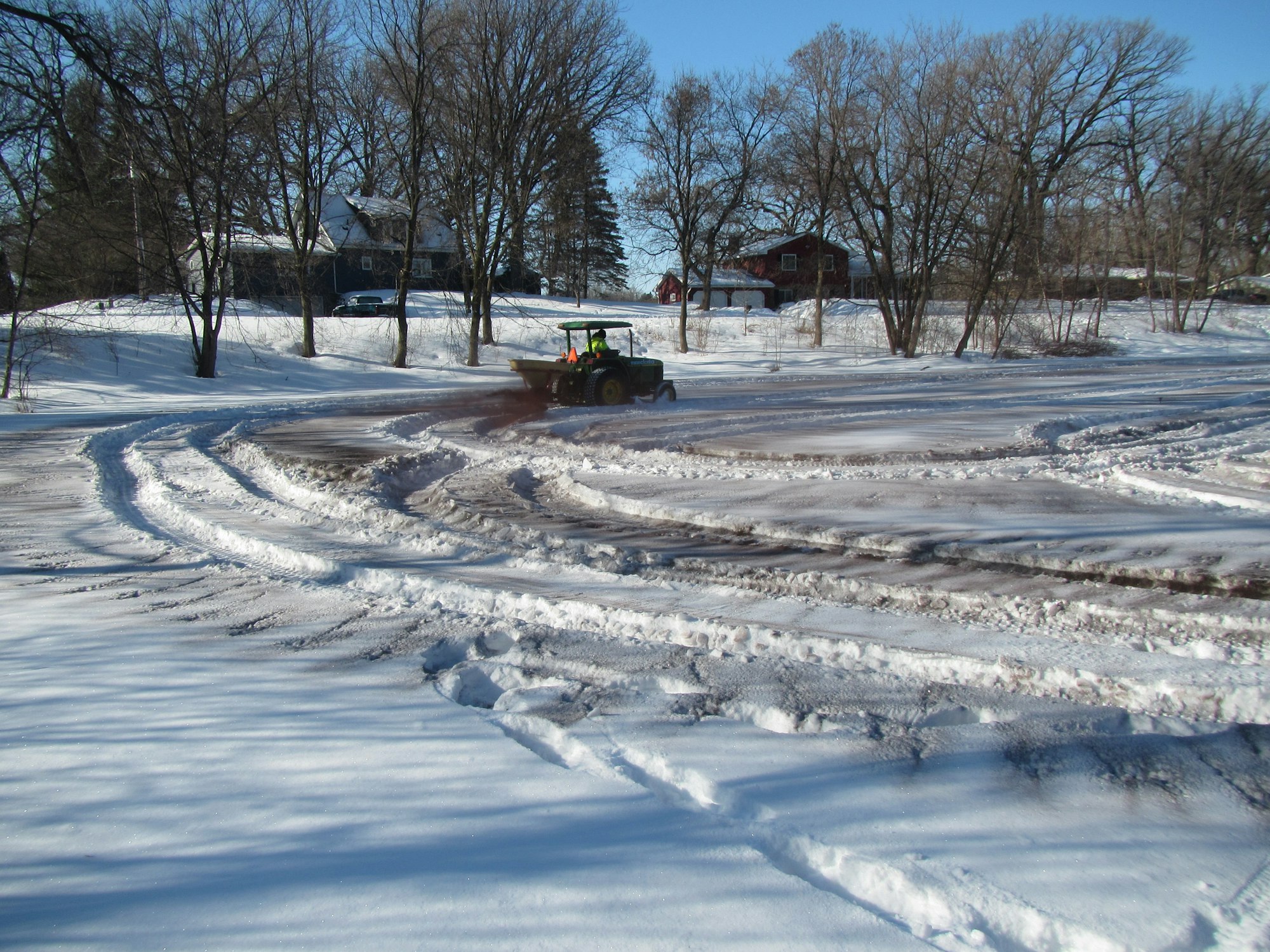 A tractor in a snowy landscape with tire tracks and houses in the background.