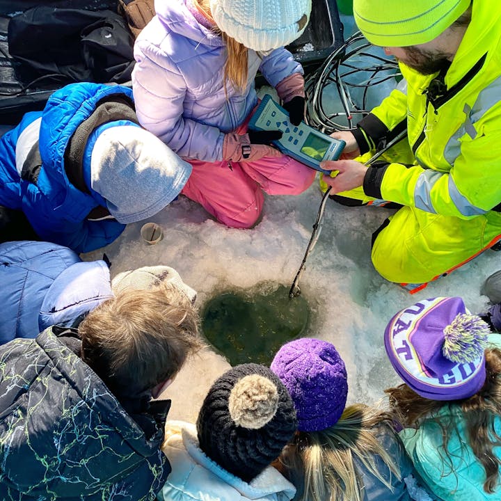 People in winter clothes examining a hole in the ice, possibly conducting some kind of scientific measurement or study.