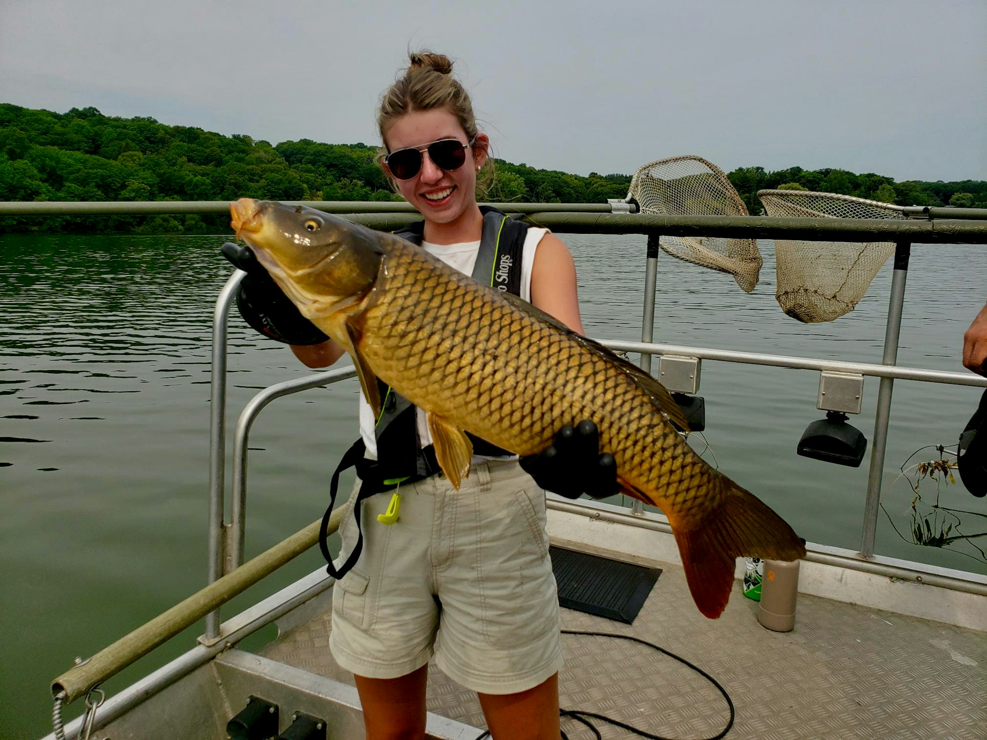A person holding a large fish on a boat, smiling, with a lake and trees in the background.