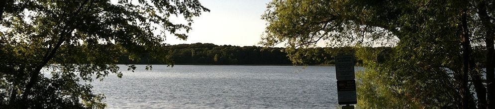A panoramic view of a serene lake with surrounding trees and a signpost to the right.