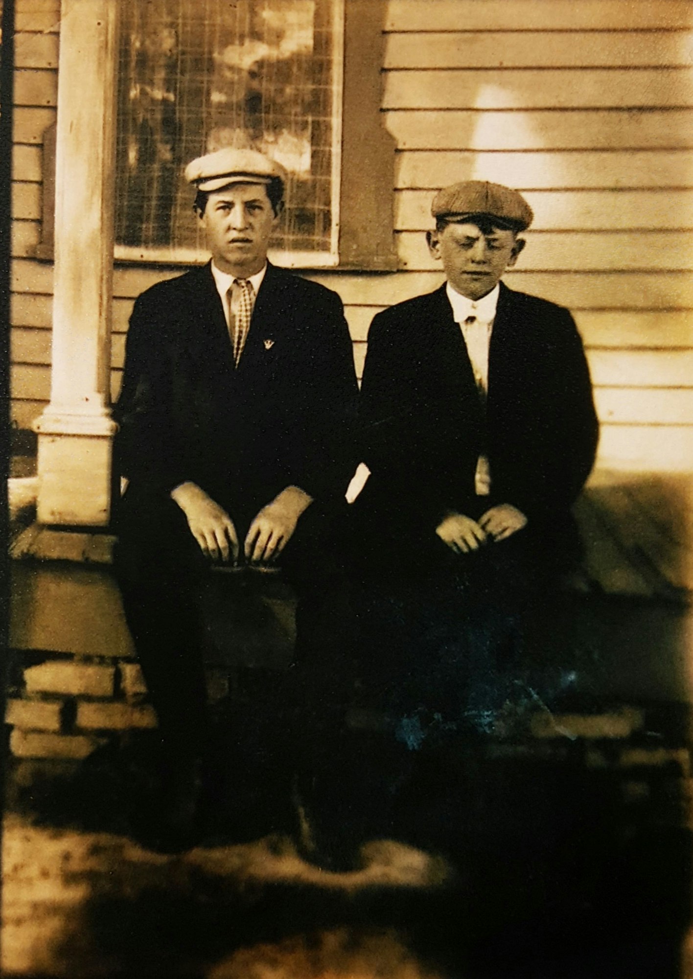 Two men sitting in front of a house, dressed in vintage clothing.