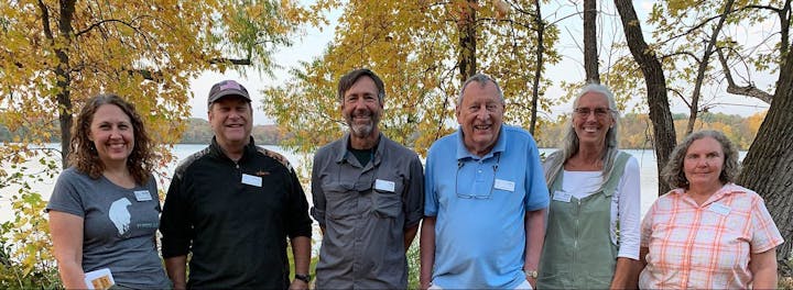 A group of six people smiling by a lake with autumn trees.