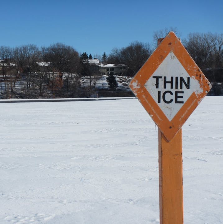 A warning sign labeled "THIN ICE" stands on a snowy surface, indicating dangerous conditions nearby. Trees and houses are in the background.