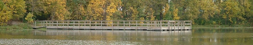 A wooden dock extends over a calm lake, surrounded by trees with autumn foliage.