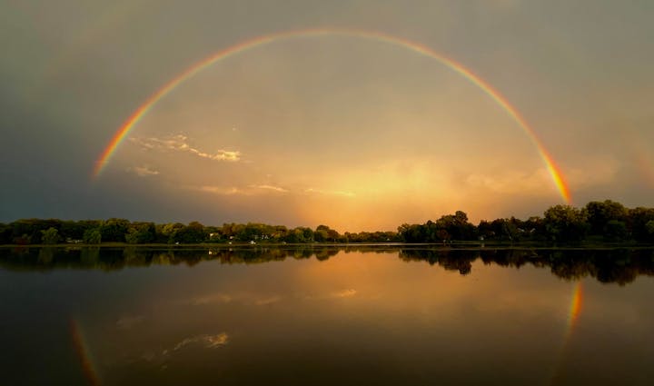 A vibrant rainbow arches over a calm lake, with the sunset sky reflected in the water.