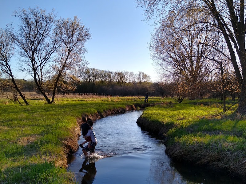 A child by a stream, trees, grassy banks, and a person in the distance, in a serene setting.