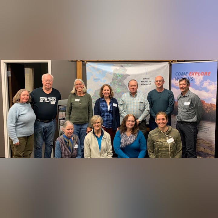 Group of people posing in front of a banner saying "Come Explore With Us" with maps in the background.