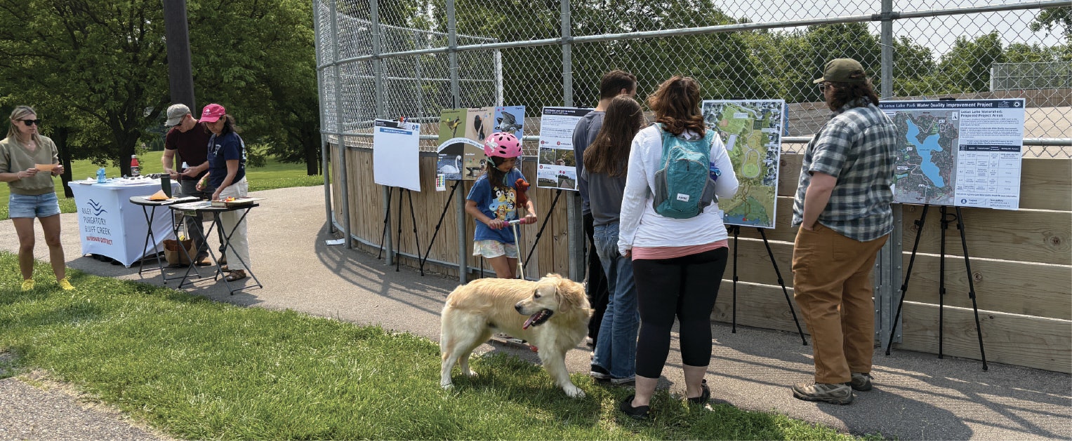 People look at informational displays near a baseball field; a dog stands nearby.