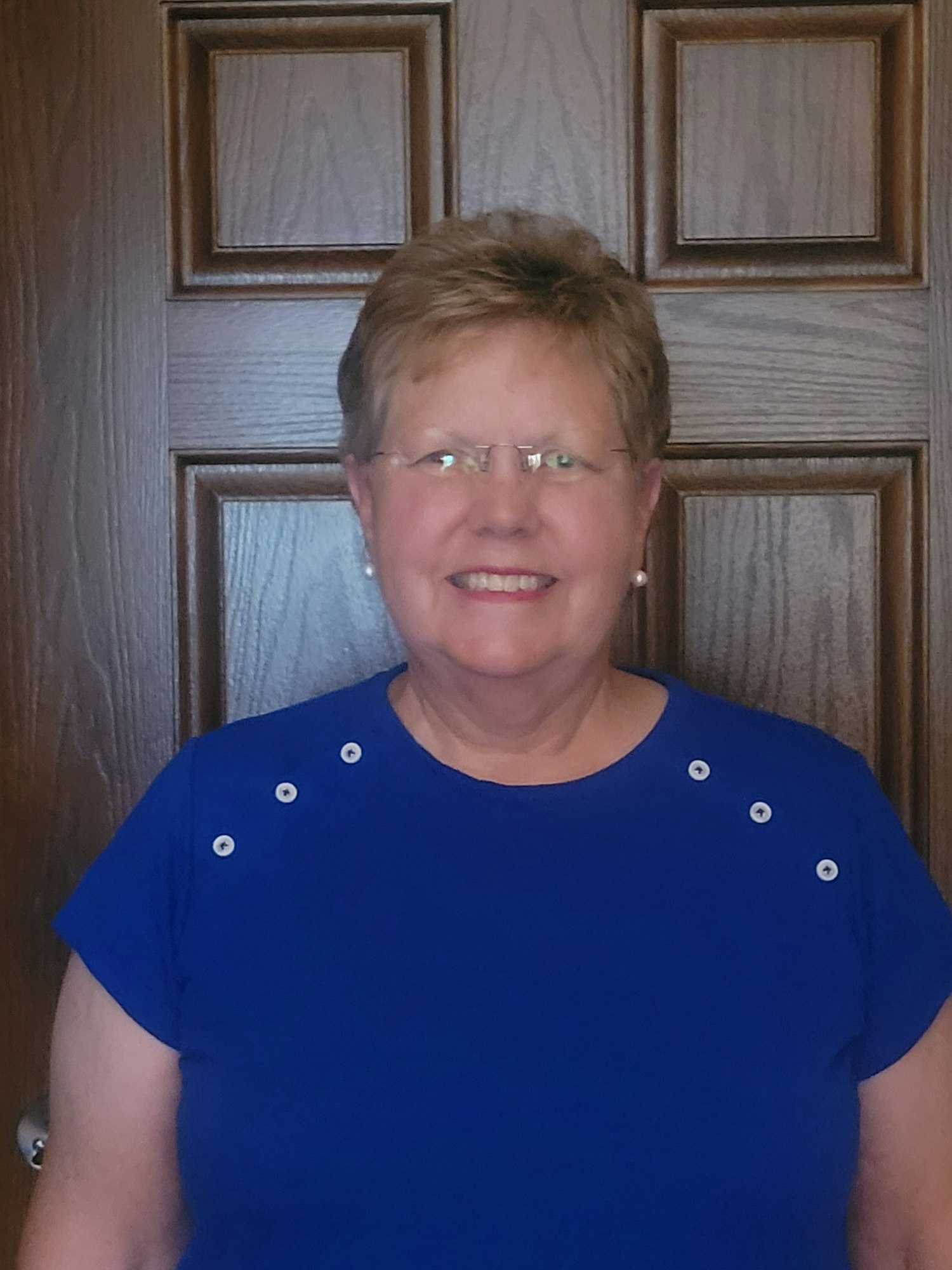 Paula Kinder smiling, wearing a blue shirt with button details, standing in front of a wooden panel door.