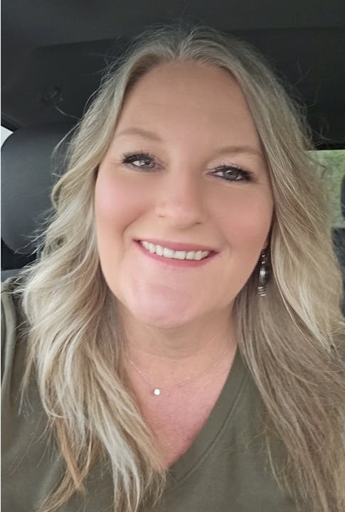 A smiling woman with long hair, wearing earrings and a pendant necklace, taken in a car.