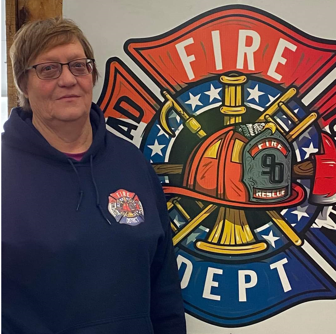 A woman wearing a hoodie stands beside a colorful fire department logo featuring a helmet and rescue tools.