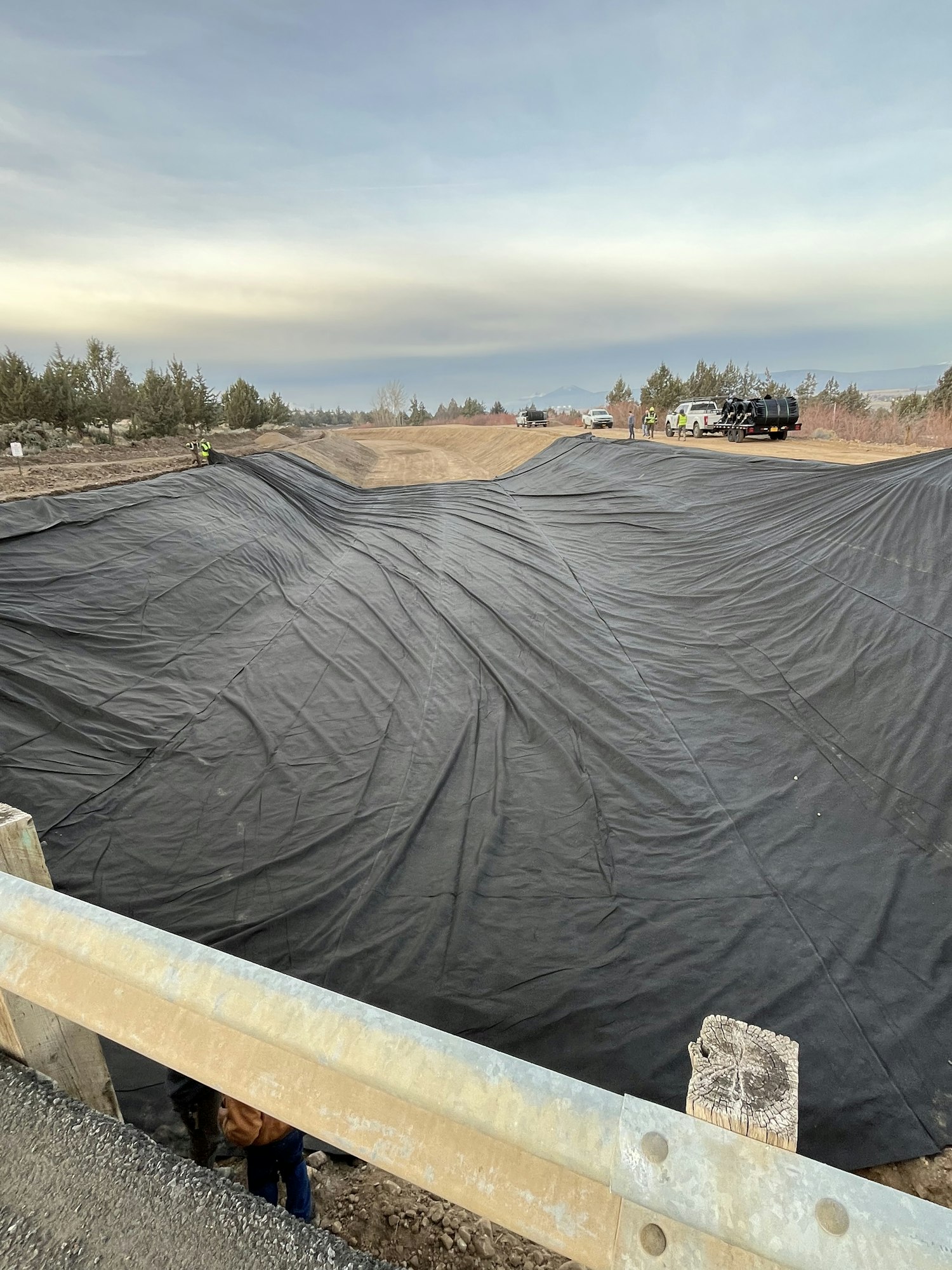 The image shows a large black tarp covering ground, with vehicles and workers visible in a construction or landscaping area.