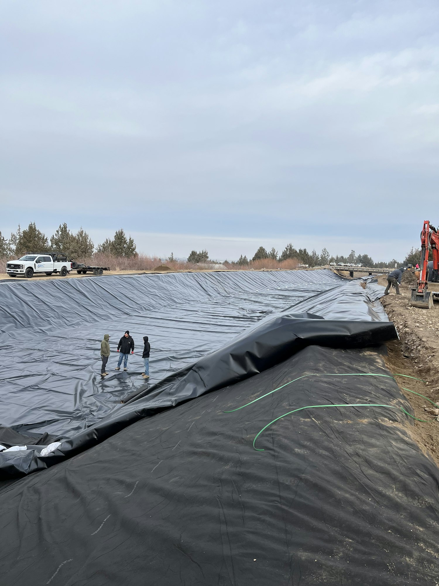 The image shows a large black tarp or liner being laid out on a construction or landscaping site with workers and equipment nearby.