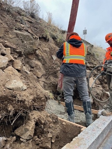 Workers in safety gear are pouring concrete near a sloped, rocky area with machinery and equipment present.