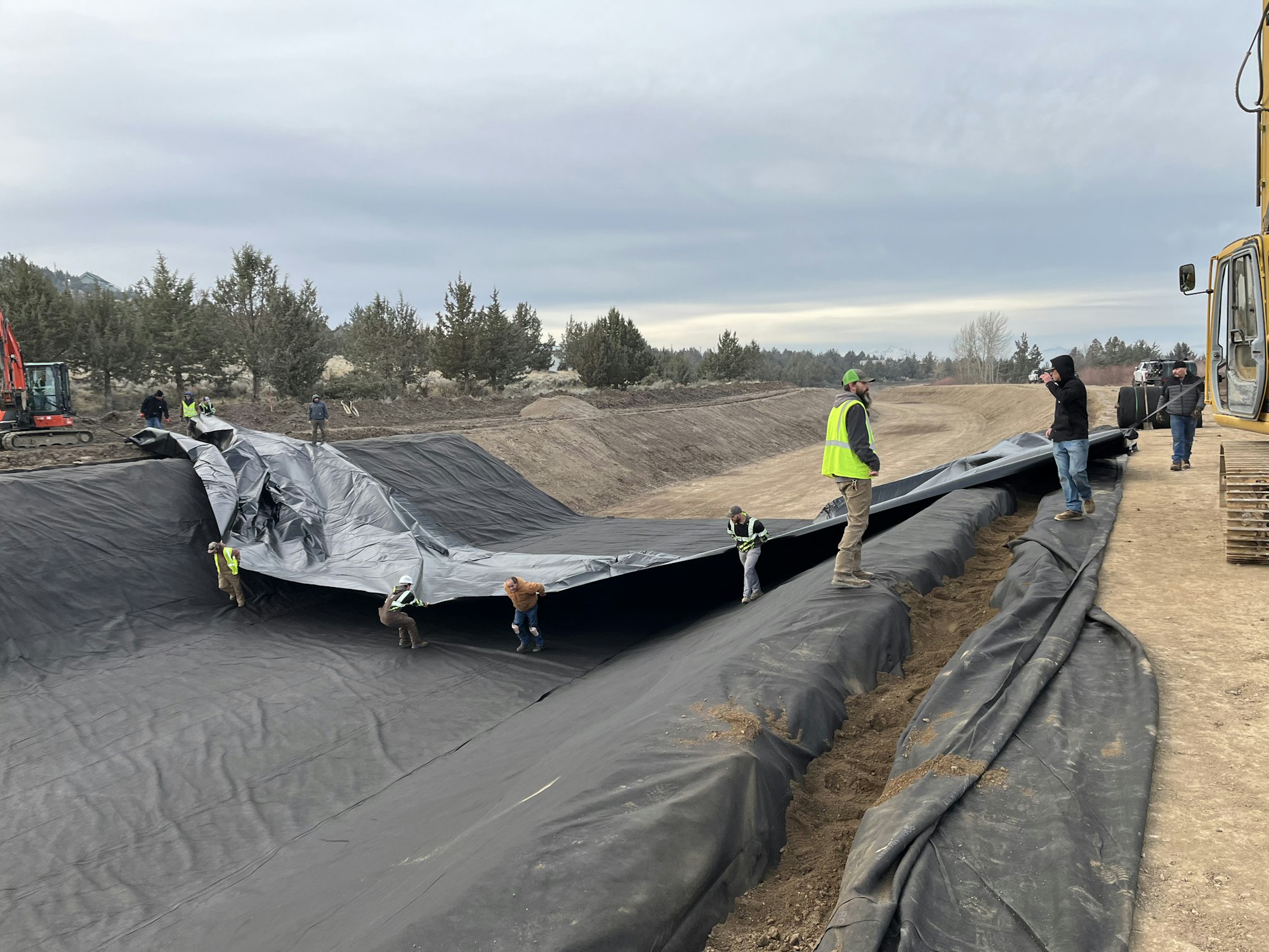Workers are unrolling large black tarps in a construction area, with machinery and trees in the background.