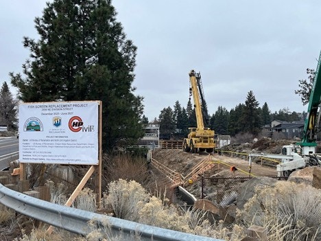 Image shows a construction site for a fish screen replacement project, featuring cranes, signage, and surrounding trees.