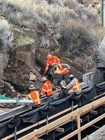 Construction workers in safety gear are working on a project near a rocky area, possibly involving excavation or repairs.