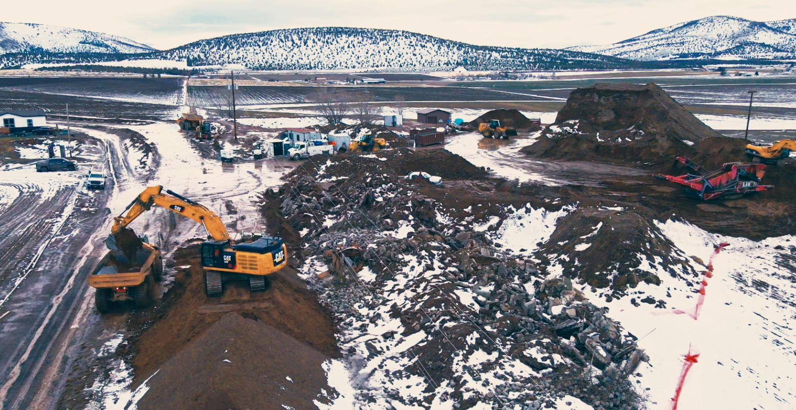 Snowy construction site with excavators and trucks working amid mounds of dirt, surrounded by mountains and fields.