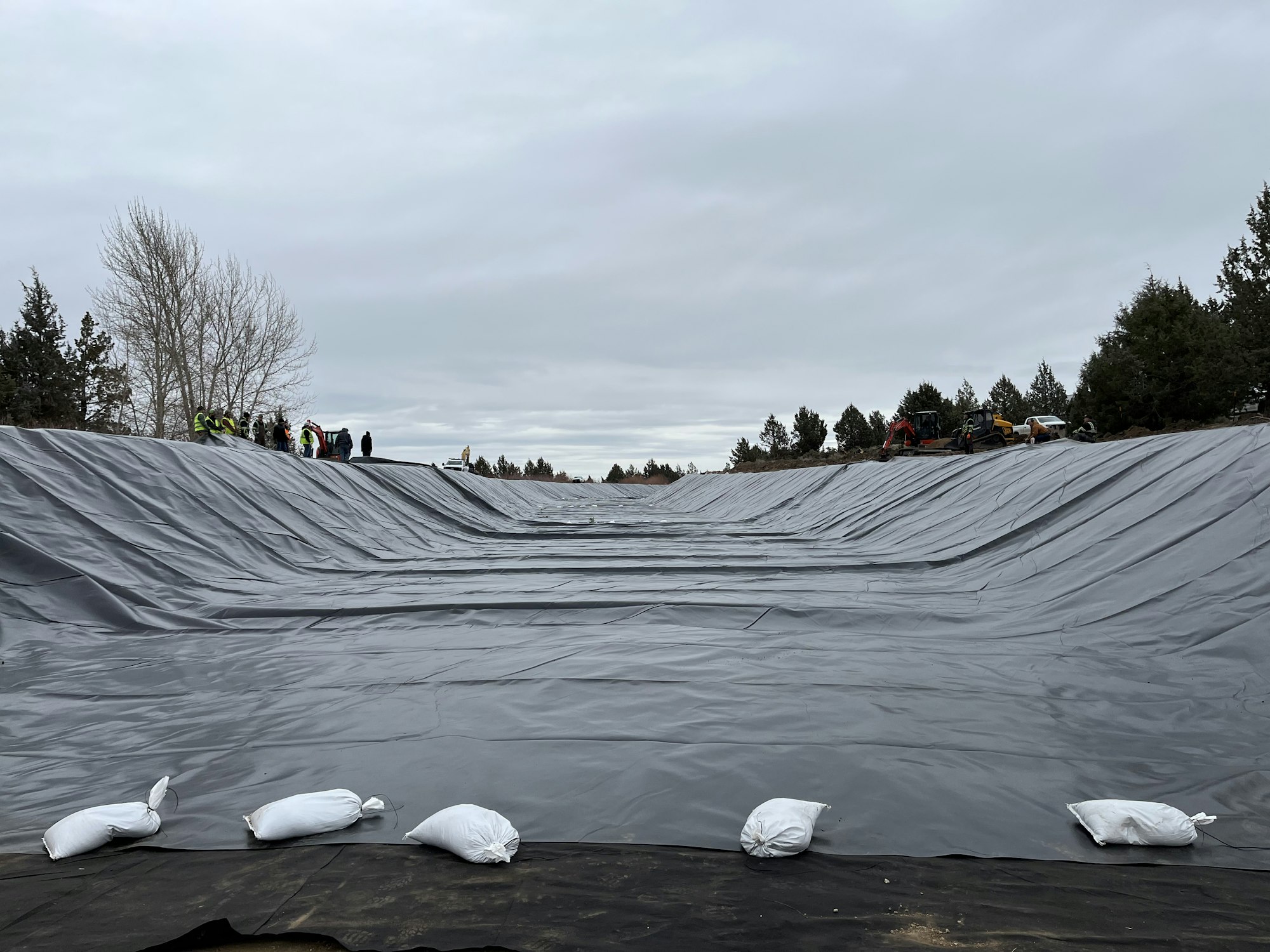 The image shows a large area covered with black tarp, workers in the background, and sandbags placed along the edge.