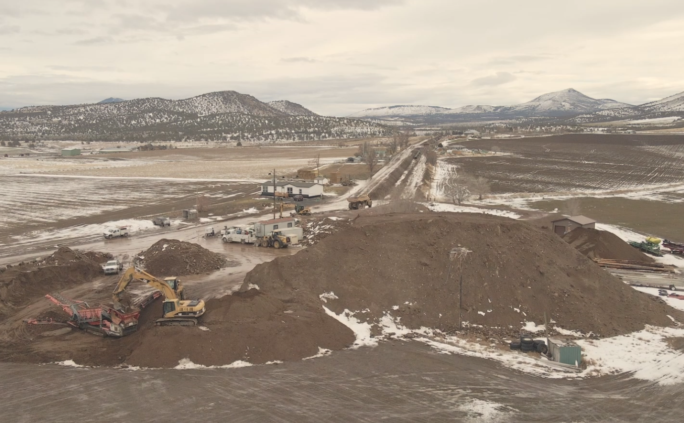 A construction site with heavy machinery, dirt piles, snow patches, and mountains in the background.