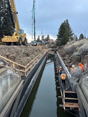A construction site with cranes and workers near a water channel, featuring wooden walkways and surrounding vegetation.