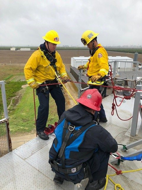 Several fighters are on top of a tower doing a training class and about to lower a firefighter over the side. 5 stories off the ground