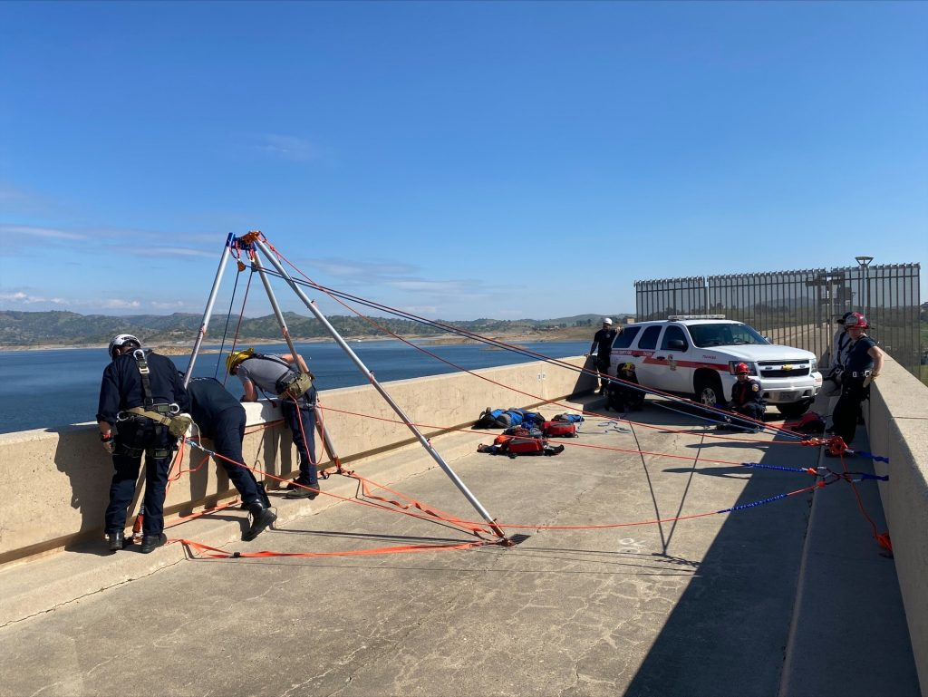 Rope rescue training on top of Millerton Lake Dam. Several fire fighters are looking over the side down the dam at the rescuer below.