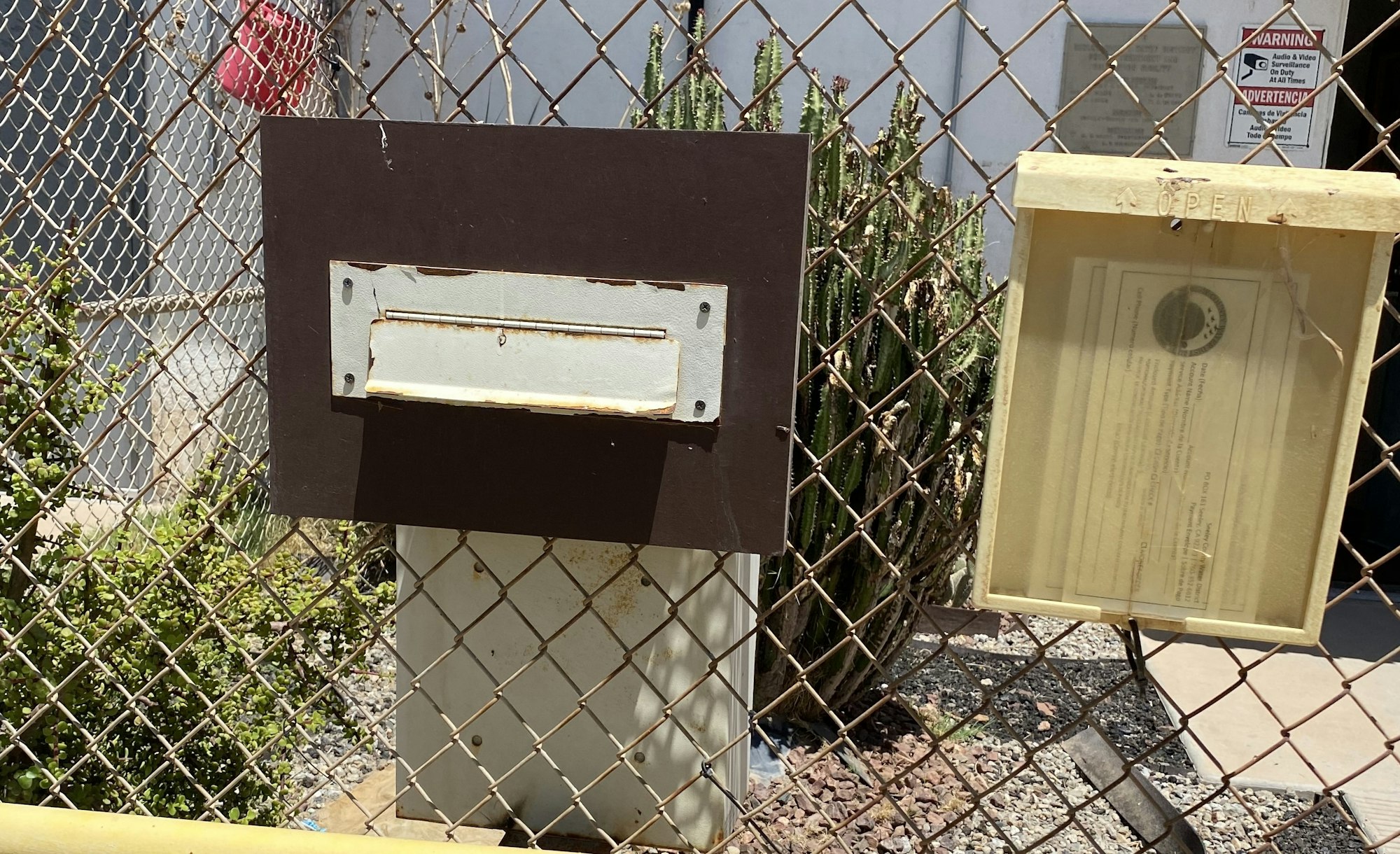 A metal mailbox slot, a warning sign, and a document in a case behind a chain-link fence, with cacti in the background.