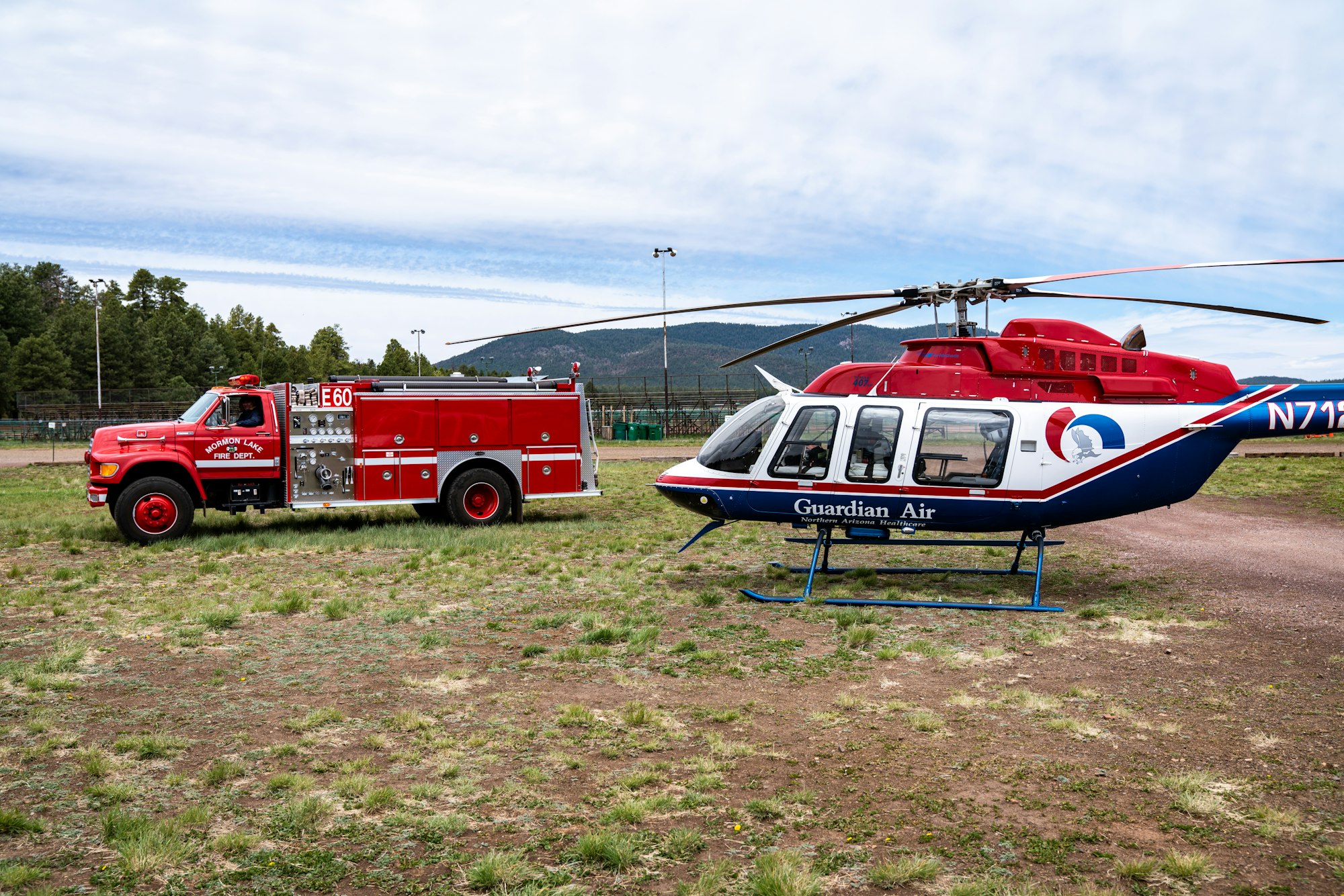 A red fire truck and a white helicopter with a medical emblem are parked on a grassy field under a cloudy sky, conveying readiness and urgency.