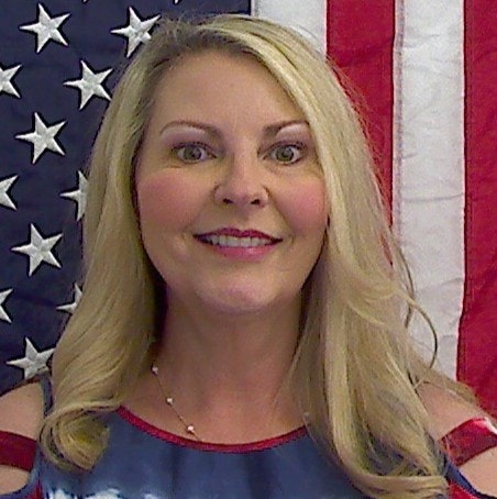 A smiling woman with blonde hair is in front of an American flag background. She's wearing a blue top with red accents.