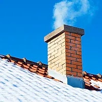 Snow-covered roof with a brick chimney emitting smoke against a clear blue sky.