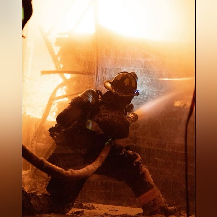 A firefighter aiming a hose at a fire, amidst smoke and glowing flames.