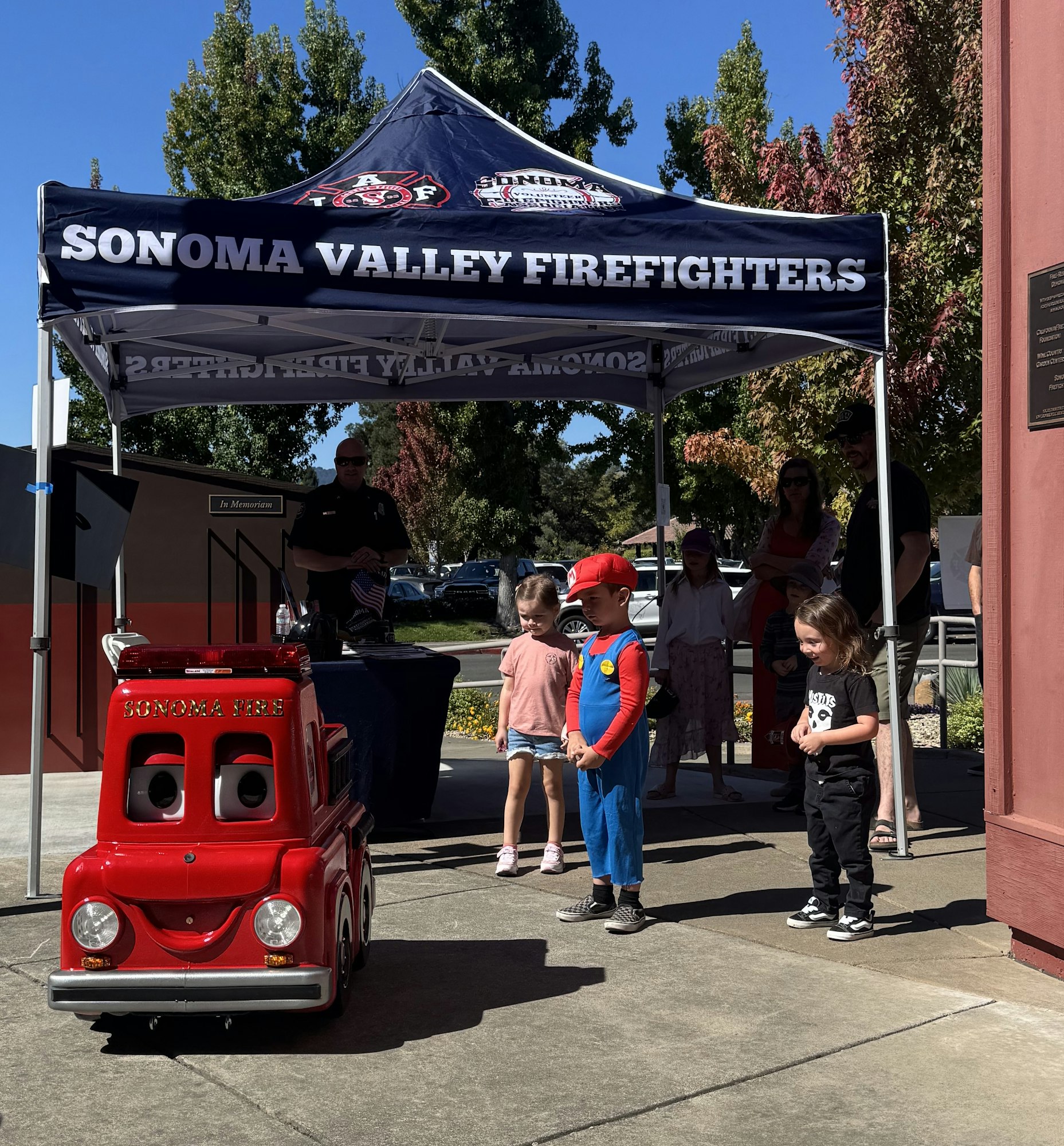 Kids stand by a small fire truck under a "Sonoma Valley Firefighters" tent, with adults and a firefighter nearby.