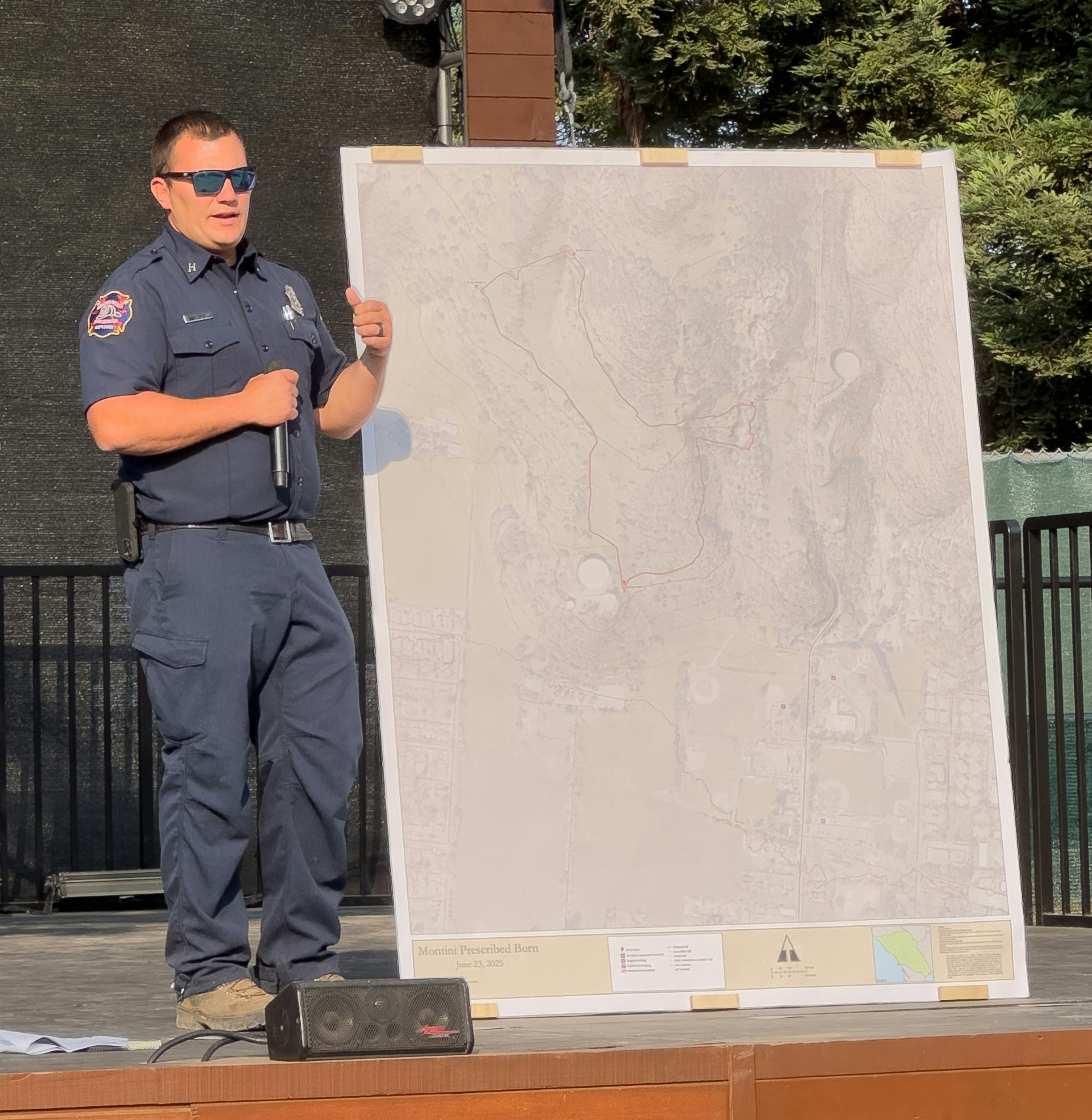 A person in a uniform holding a microphone stands beside a large map on a board outdoors.
