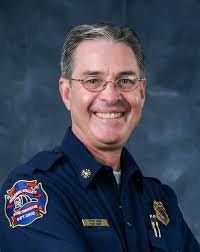 A smiling man in a firefighter uniform with glasses, posing in front of a plain background.