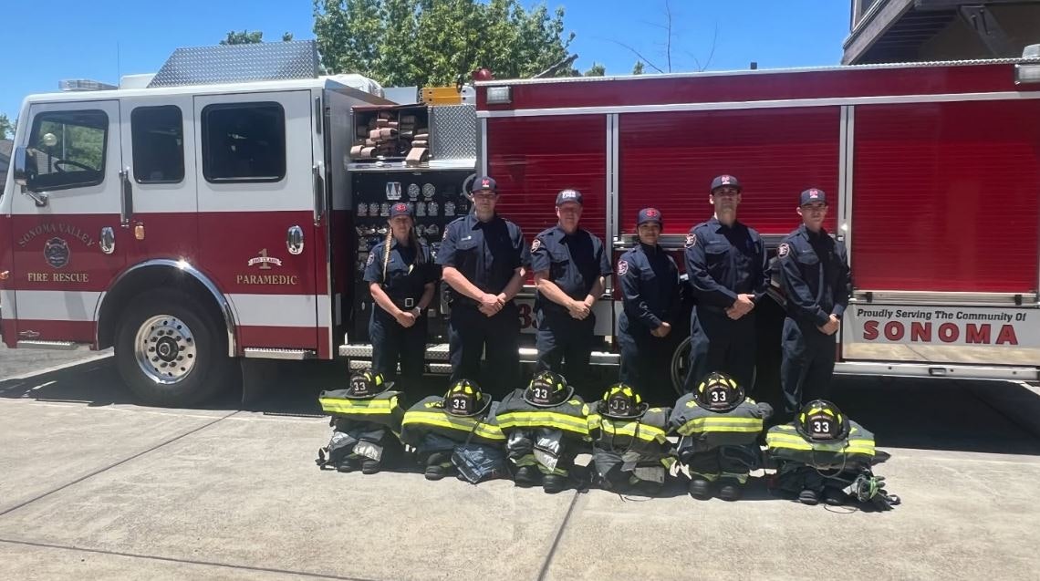 A group of firefighters stands in front of a fire truck, with their gear laid out on the ground in front of them.