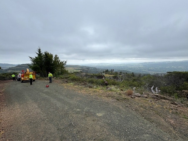 A gravel road in a hilly area with workers in bright clothing, vehicles, and a cloudy sky in the background.