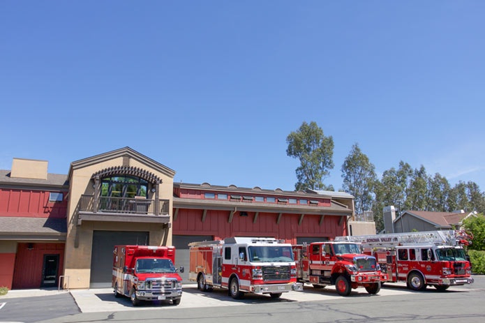 A fire station with three red fire trucks and an ambulance parked outside under a blue sky.