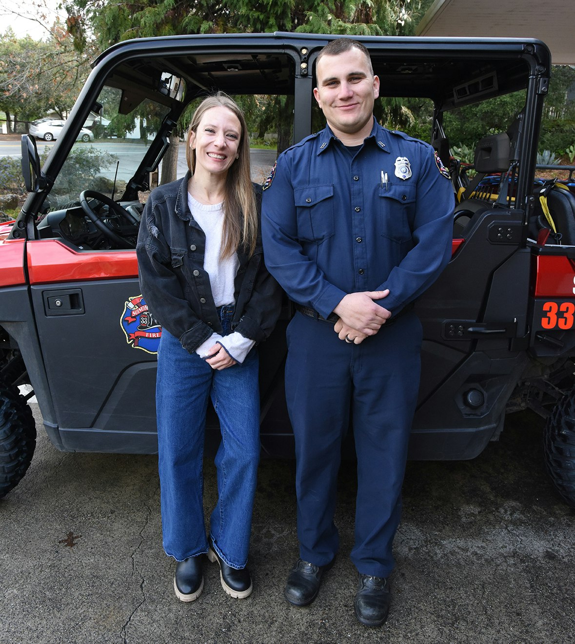 A woman and a man in uniform pose together in front of a fire department vehicle. Both are smiling.