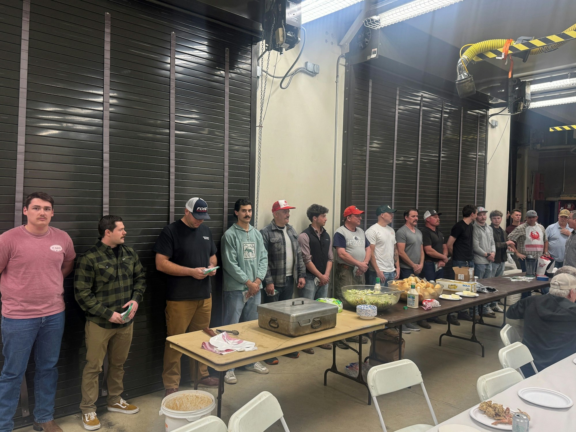 A group of men stands in a line by a table with food, possibly at a gathering or event in a community space.
