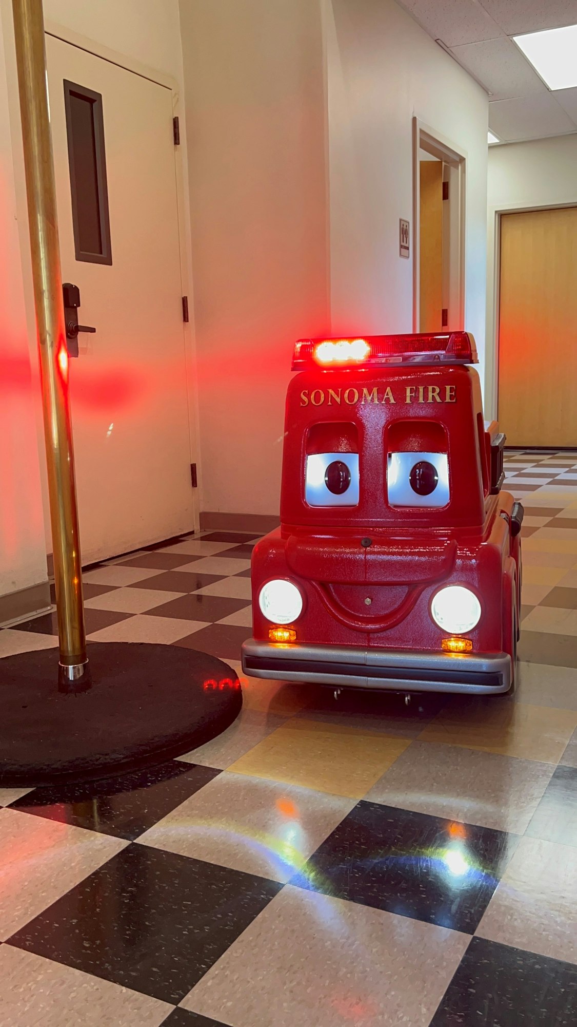 A cartoon-like fire truck with "SONOMA FIRE" on it, lights flashing, beside a fire pole in a hallway.